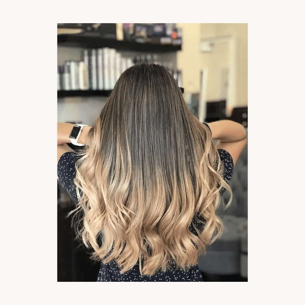 Woman with long, wavy ombre hair facing away, in a salon setting.