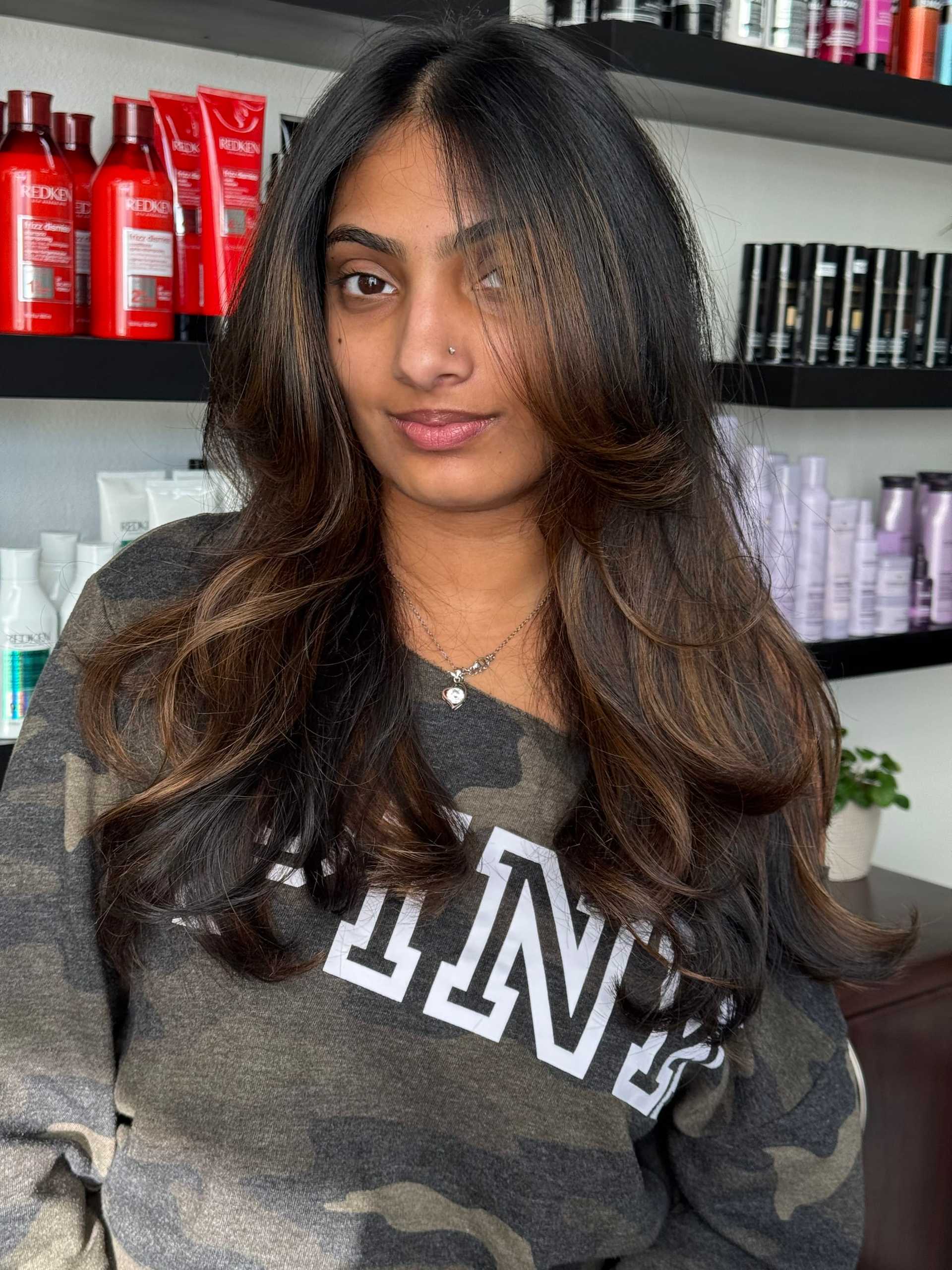Young woman with long wavy hair in salon, wearing a camo-patterned "Pink" sweater.